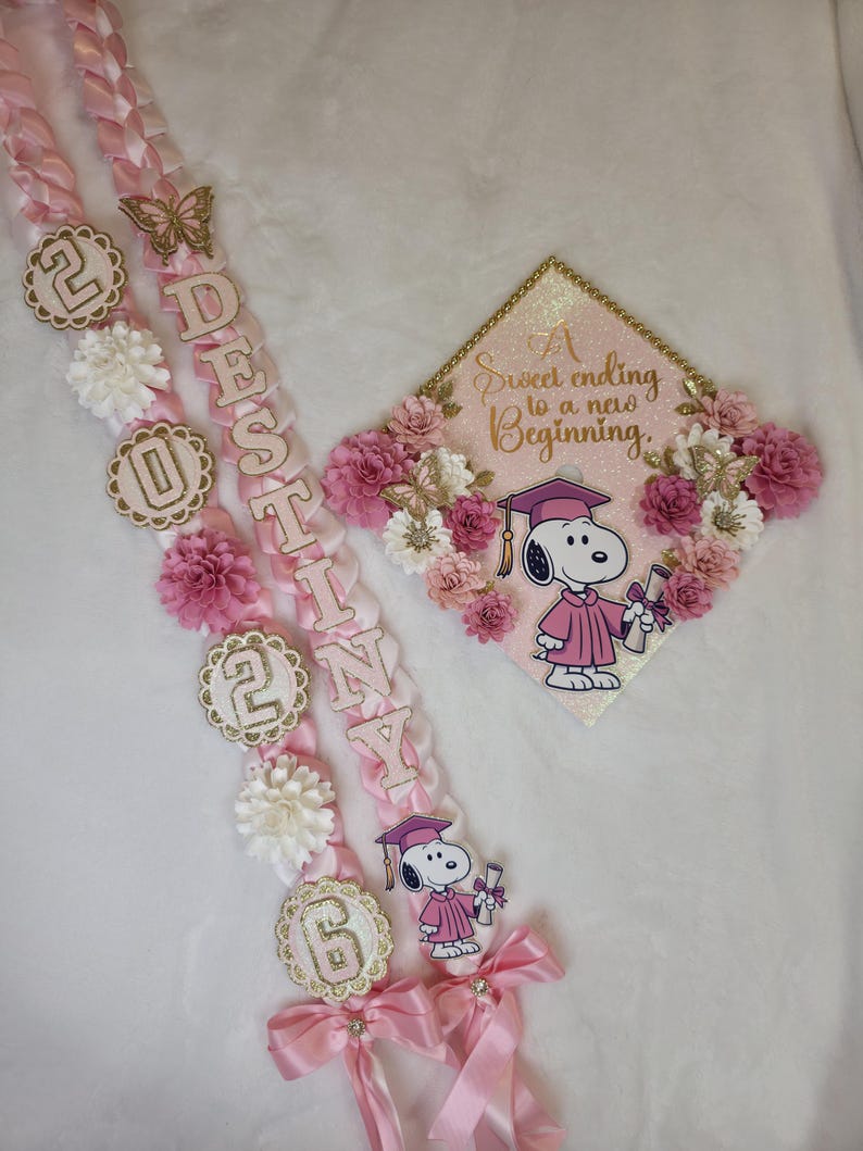 Puede incluir: Conjunto de lei y gorra de graduaci&oacute;n rosa y dorado. El lei deletrea el nombre "Destiny" e incluye el a&ntilde;o "2026". La gorra presenta a Snoopy con toga de graduaci&oacute;n y el texto "A Sweet ending to a new Beginning."