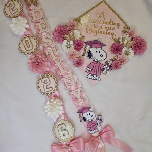 Puede incluir: Guirnaldas y gorro de graduaci&oacute;n rosas y blancos con gr&aacute;ficos de Snoopy. Las guirnaldas deletrean el nombre "Destiny" y presentan los n&uacute;meros "2026". El gorro tiene el texto "A sweet ending to a new beginning."