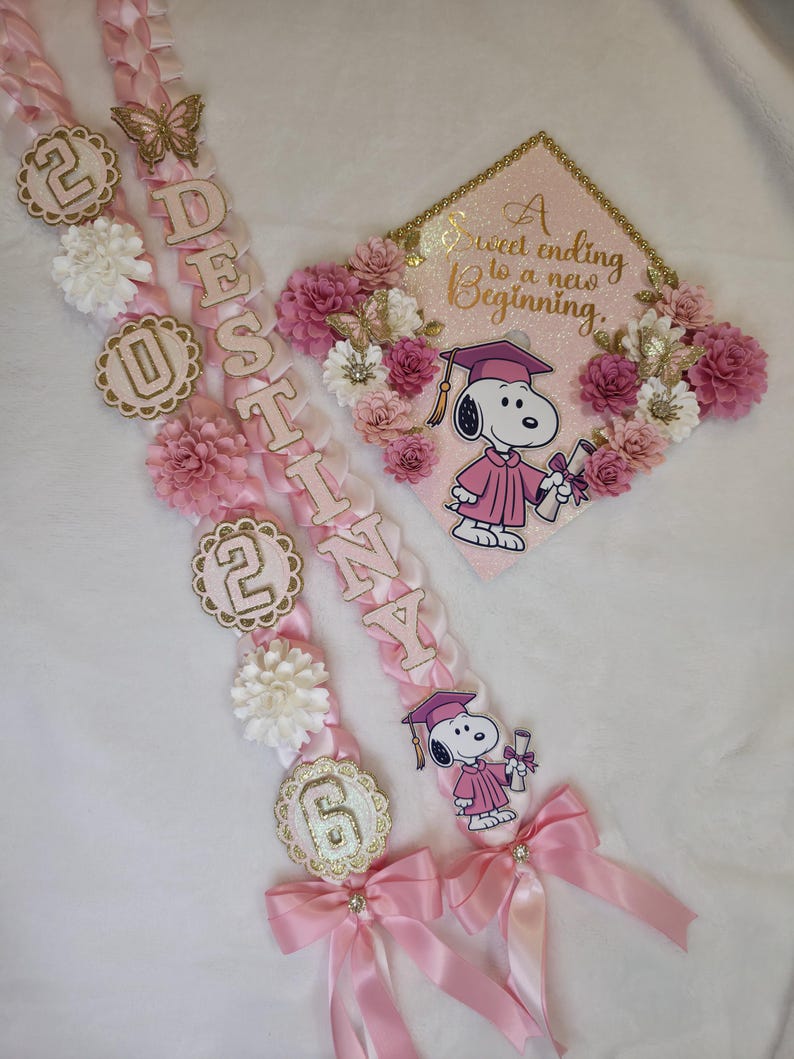 Puede incluir: Guirnaldas y gorro de graduaci&oacute;n rosas y blancos con gr&aacute;ficos de Snoopy. Las guirnaldas deletrean el nombre "Destiny" y presentan los n&uacute;meros "2026". El gorro tiene el texto "A sweet ending to a new beginning."