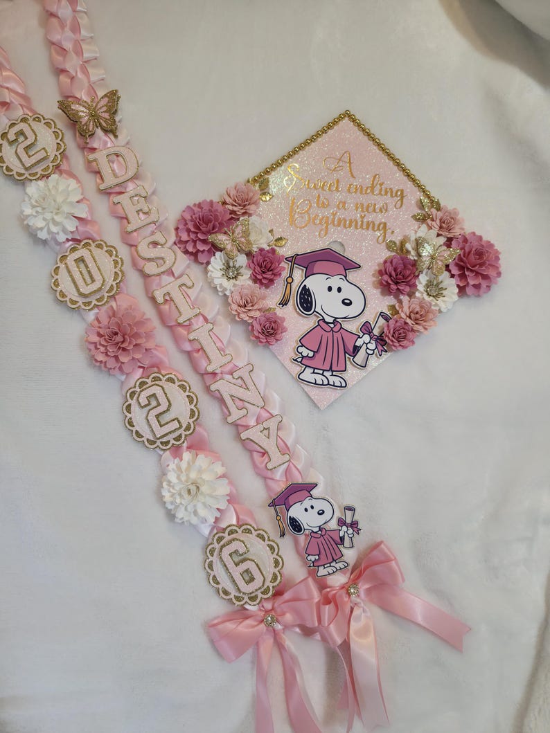 Puede incluir: Guirnalda y gorra de graduaci&oacute;n rosas con Snoopy con toga de graduaci&oacute;n. La guirnalda tiene el nombre "Destina" y el a&ntilde;o "2026" en letras doradas. La gorra tiene el texto "A sweet ending to a new beginning".