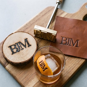 Op de afbeelding: Een messing brandijzer met de letters BJM, een ronde houten schijf met het BJM-merk, een vierkant bruin leren stuk met het BJM-merk en een glas met amberkleurige vloeistof en een sinaasappelschil met het BJM-merk op een houten snijplank.