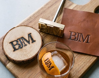 May include: A brass branding iron with the letters BJM, a round wood slice with the BJM brand, a square brown leather piece with the BJM brand, and a glass of amber liquid with an orange peel featuring the BJM brand on a wood cutting board.