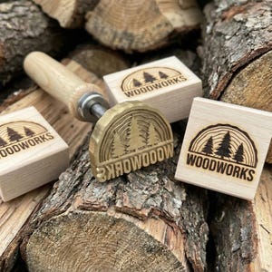 Op de afbeelding: Een metalen brandstempel met een houten handvat en een halfronde kop met een dennenboomontwerp en de tekst WOODWORKS, vergezeld van twee vierkante houten blokken die elk zijn gestempeld met hetzelfde dennenboom- en WOODWORKS-logo.