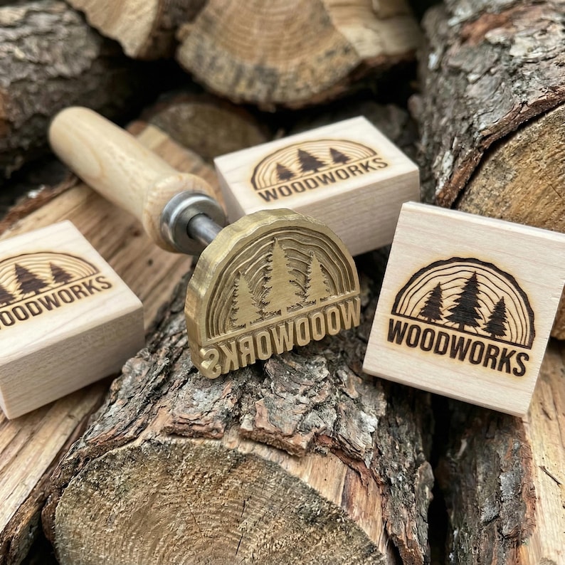 Op de afbeelding: Een metalen brandstempel met een houten handvat en een halfronde kop met een dennenboomontwerp en de tekst WOODWORKS, vergezeld van twee vierkante houten blokken die elk zijn gestempeld met hetzelfde dennenboom- en WOODWORKS-logo.