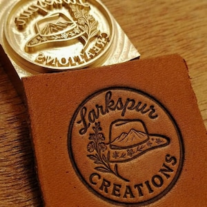 Op de afbeelding: Een ronde messing stempel met een gravure van een cowboyhoed en bloemmotieven, geplaatst boven een vierkant stuk bruin leer dat is bedrukt met hetzelfde ontwerp en de tekst Larkspur CREATIONS in een cirkelvormige lay-out.