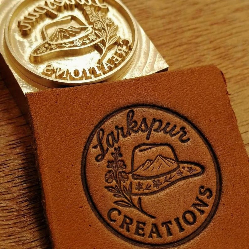Op de afbeelding: Een ronde messing stempel met een gravure van een cowboyhoed en bloemmotieven, geplaatst boven een vierkant stuk bruin leer dat is bedrukt met hetzelfde ontwerp en de tekst Larkspur CREATIONS in een cirkelvormige lay-out.