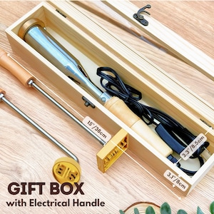 Op de afbeelding: Een elektrisch brandijzer met een houten handvat en een metalen verwarmingselement, vergezeld van een aparte ronde messing stempel met de letters SB. Het gereedschap zit in een lichthouten geschenkdoos van 38 cm bij 8,5 cm bij 8 cm.