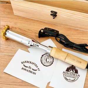 Op de afbeelding: Een metalen brandstempel met een houten handvat, een goudkleurige knop en een zwart netsnoer met een aan-uit-schakelaar. Het handvat is gemarkeerd met 110V/350W. Drie witte kaarten tonen de logo's: Handcrafted By Jonathan, een letter C en Jonathan Woodworks.