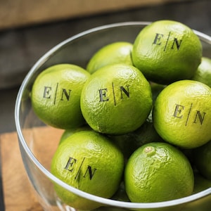Op de afbeelding: Een doorzichtige glazen kom gevuld met heldergroene limoenen. Verschillende limoenen zijn bedrukt met een zwart inktmonogram met de letters E en N gescheiden door een verticale lijn. De limoenen hebben een getextureerde, bobbelige schil.