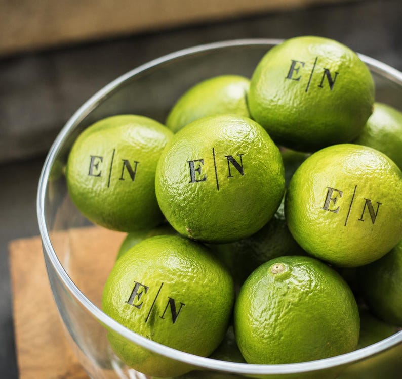 Op de afbeelding: Een doorzichtige glazen kom gevuld met heldergroene limoenen. Verschillende limoenen zijn bedrukt met een zwart inktmonogram met de letters E en N gescheiden door een verticale lijn. De limoenen hebben een getextureerde, bobbelige schil.