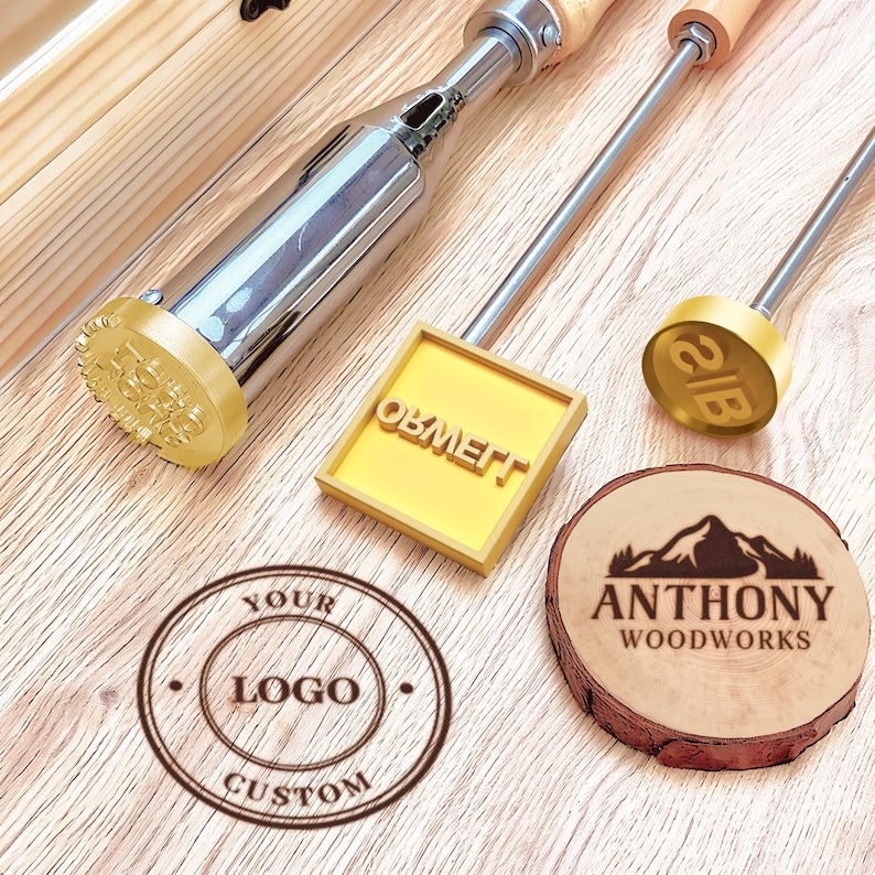 Op de afbeelding: Drie metalen brandijzers met houten handvatten, waaronder een ronde kop, een vierkante kop met de tekst ORWELL en een kleine ronde kop met de letters IB. Een cirkelvormige logostempel met YOUR LOGO CUSTOM en een houten schijf met ANTHONY WOODWORKS.