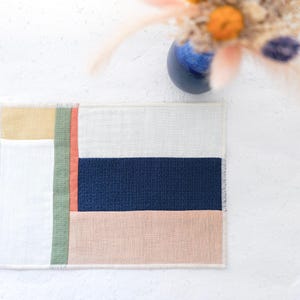 Könnte beinhalten: Rechteckiger Textilartikel mit Patchwork-Design. Das Design zeigt Blöcke in Weiß, Marineblau, Pfirsich, Gelb und Grün. Der Textilartikel ist wahrscheinlich ein Platzset oder ein kleiner Teppich mit strukturierter Optik und Fransen.