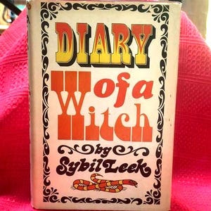May include: A vintage book titled "Diary of a Witch" by Sybil Leek. The book cover features bold orange and yellow lettering against a cream background, with decorative black scrollwork and a snake illustration. The book is set against a pink background.