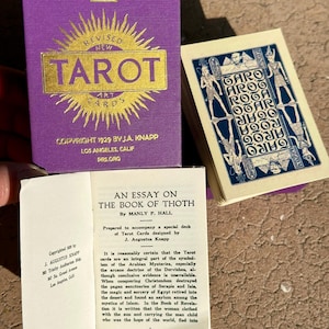 May include: A vintage Tarot card set with a purple box featuring gold lettering. The box reads "REVISED NEW TAROT ART CARDS" and "COPYRIGHT 1929 BY J.A. KNAPP". A booklet titled "AN ESSAY ON THE BOOK OF THOTH" is also visible.
