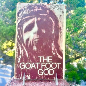 May include: A rectangular book titled The Goat Foot God by Dion Fortune. The cover features a dark brown, abstract, textured illustration of a face on a cream-colored background. The title and author name are printed in white text.