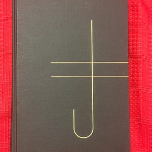 May include: A dark gray hardcover book with a gold-colored geometric design on the cover. The design consists of a vertical line intersected by a horizontal line and a curved line. The book is on a red patterned surface.