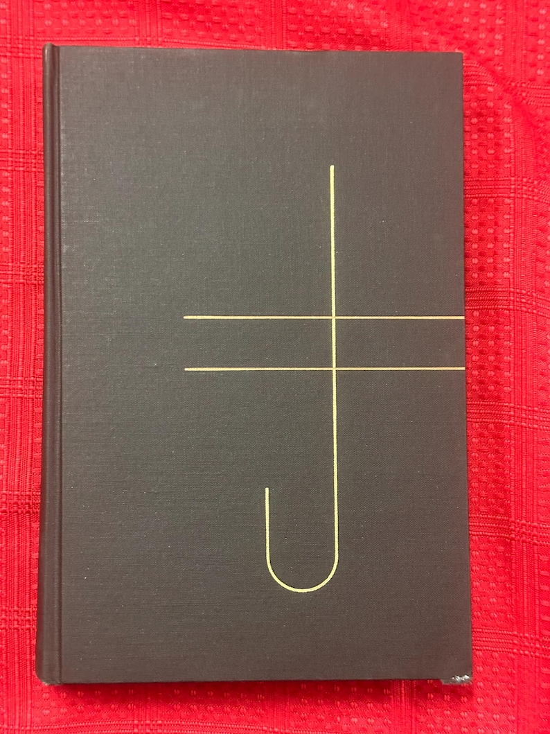 May include: A dark gray hardcover book with a gold-colored geometric design on the cover. The design consists of a vertical line intersected by a horizontal line and a curved line. The book is on a red patterned surface.