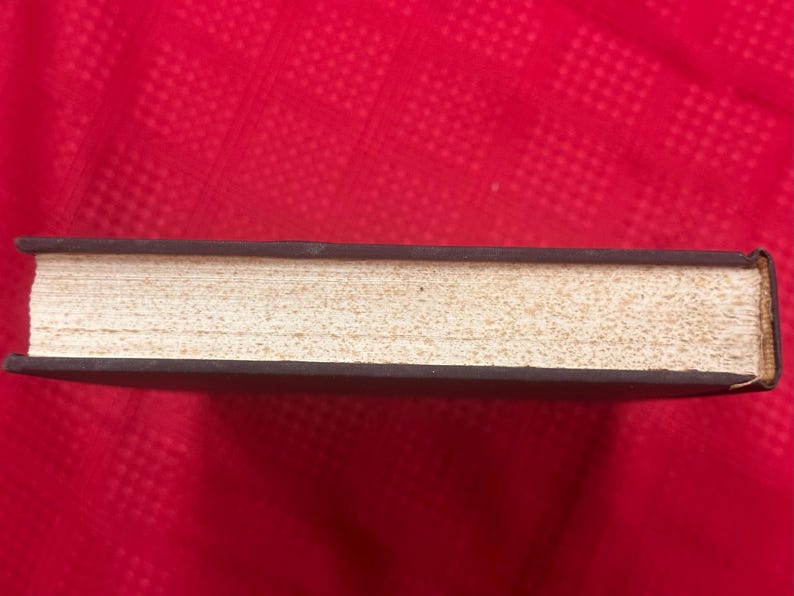 May include: A closed book with a dark brown cover and aged, off-white pages. The book's spine is visible, and the pages have a textured appearance. The book is set against a red background.