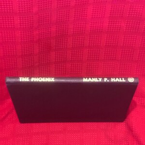 May include: A book titled "THE PHOENIX" by Manly P. Hall. The book has a dark purple cover with white lettering. The book is set against a red textured background.