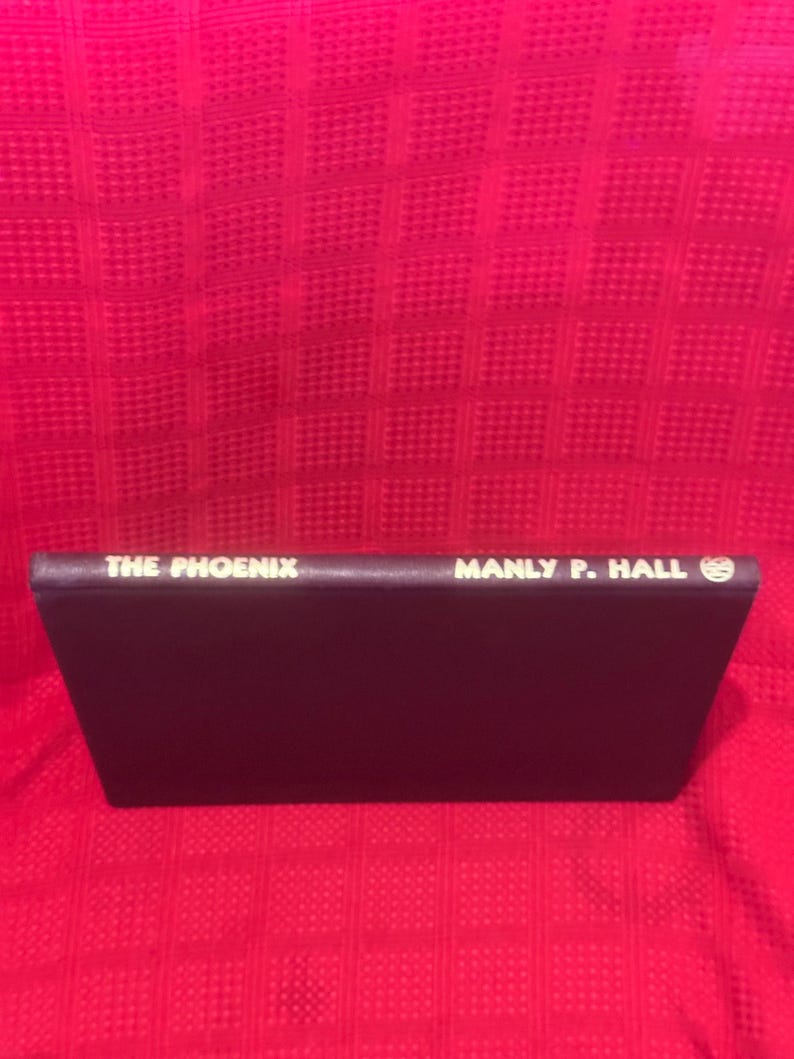 May include: A book titled "THE PHOENIX" by Manly P. Hall. The book has a dark purple cover with white lettering. The book is set against a red textured background.