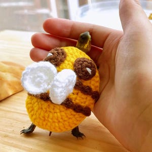 Può includere: Un'ape all'uncinetto fatta a mano con dettagli gialli, marroni e bianchi. L'ape ha strisce marroni, ali bianche e piccole zampe. L'ape è tenuta in una mano, mostrando gli intricati dettagli dell'artigianato.