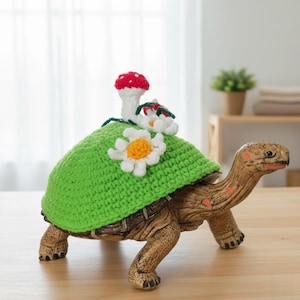 Puede incluir: Figura decorativa de tortuga con una cubierta de caparazón de ganchillo verde, adornada con una margarita blanca y una seta roja y blanca. La tortuga es marrón con manchas naranjas, sobre una superficie de madera.