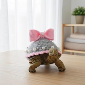 Puede incluir: Figura decorativa de tortuga con un sombrero de ganchillo gris con un lazo rosa y detalles de perlas. El sombrero tiene un borde rosa y está adornado con perlas de imitación. La tortuga es marrón y verde y está sobre una superficie de madera.