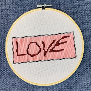 May include: A cross-stitch embroidery hoop featuring the word "LOVE" in dark brown on a red background. The embroidery is framed by a light yellow wooden hoop and set against a white fabric.