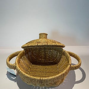 May include: A woven wicker basket with a lid and two handles. The basket is light brown and has a natural finish.