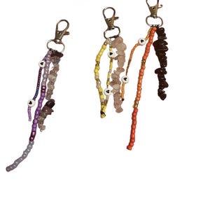 May include: Three keychains with silver-colored clasps. Each keychain features a different design of beads and small stones in various colors, including purple, yellow, orange, and brown. Some strands have small white heart-shaped beads.