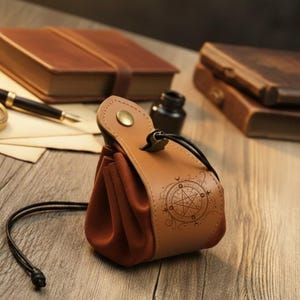 Può includere: Un sacchetto in pelle marrone con chiusura a coulisse e un design decorativo a pentagramma. Il sacchetto ha un interno simile alla pelle scamosciata e una chiusura a scatto. Altri oggetti sullo sfondo includono libri rilegati in pelle e una penna.