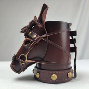 May include: A brown leather horse head shaped cup holder with brass accents. The cup holder has a strap that goes around the neck of the horse head.