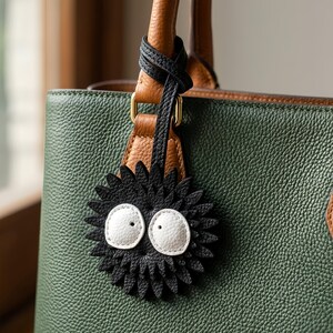 Può includere: Una borsa in pelle verde con dettagli in pelle marrone e un cinturino in pelle nera. Un ciondolo nero e appuntito con occhi bianchi pende dal manico. Il ciondolo ha un aspetto da cartone animato.