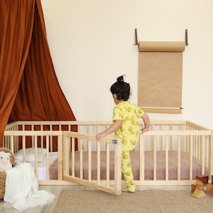 Op de afbeelding: Een licht houten babybed met een hekje, een bruine luifel en een kind in gele pyjama's. Een rieten mand met knuffels en een witte deken staat naast het bed. Een rol papier hangt aan de muur.
