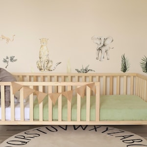 Op de afbeelding: Een houten ledikant met een lichtgroene matras en een grijze knuffelolifant. Het ledikant heeft een slinger van bruine driehoeken. De muur achter het ledikant heeft dierenthema muurstickers, waaronder een cheetah en een olifant.