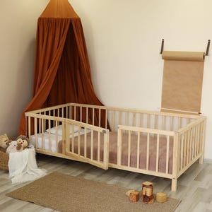 May include: A light wood toddler bed with a side gate, a brown canopy, and a small rug. A basket of stuffed animals and a bookshelf with children's books are also visible. The bed has a mattress and a pink blanket.