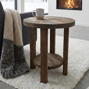 May include: A rustic, round wooden side table with a lower shelf. The table has a weathered, natural finish and is supported by four sturdy legs. A mug sits on top. A cozy blanket and chair are nearby.
