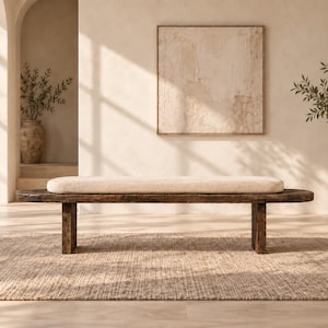 May include: A long, low-profile bench featuring a dark, weathered wooden base with a natural grain texture and a thick, rectangular off-white upholstered cushion that spans the length of the seat, supported by two sturdy, block-style wooden legs.