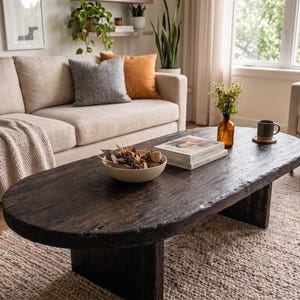 May include: An oval coffee table made of dark, weathered wood with a thick, uneven edge and a sturdy pedestal base. The table surface holds a beige bowl filled with dried botanicals, a stack of two books, a small amber glass vase, and a mug.
