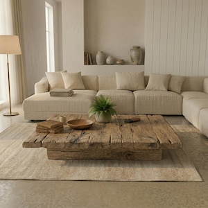Op de afbeelding: Een lichte woonkamer met een grote hoekbank in neutrale tinten, een rustieke houten salontafel en een beige tapijt. De kamer wordt overspoeld met natuurlijk licht, wat een warme en uitnodigende sfeer creëert. Decoratieve elementen zijn aanwezig.