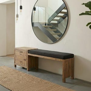 Puede incluir: Un banco de madera con un armario empotrado y un cojín gris oscuro. Un gran espejo redondo cuelga de la pared sobre el banco. Una alfombra de yute tejida y plantas en macetas completan la decoración.