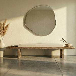 May include: A neutral-toned interior with a large, organic-shaped mirror as a focal point. A wooden bench with a light-coloured cushion is positioned below. A vase with dried grasses and a small bowl complete the scene.