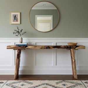 May include: A natural wood console table with a live edge design and angled legs. A round gold mirror hangs above the table, and a small framed artwork is to the left. A decorative rug is in front of the table. The table is approximately 150 cm long.