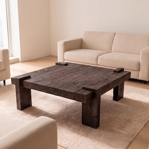 Puede incluir: Una mesa de centro cuadrada construida con madera recuperada de color marrón oscuro y aspecto desgastado. La mesa presenta una superficie gruesa y texturizada con vetas visibles y cuatro patas robustas en forma de bloque unidas a las esquinas.