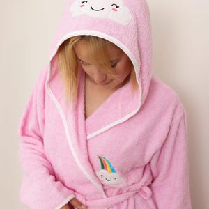 Pode incluir: Robe rosa com capuz com acabamento branco e um desenho de nuvem no capuz. O roupão apresenta um aplique de arco-íris e nuvem na frente. O roupão tem cinto e bolsos.