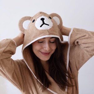 Pode incluir: Um roupão marrom com capuz, acabamento branco e um rosto de urso bordado no capuz. O roupão é feito de um material macio e felpudo.