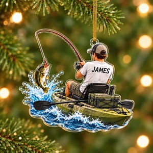May include: A clear acrylic Christmas ornament featuring a person in a kayak fishing for a bass. The person wears a white t-shirt with the name JAMES printed on the back, a grey cap, and shorts. A fishing rod is arched with a hooked fish splashing.