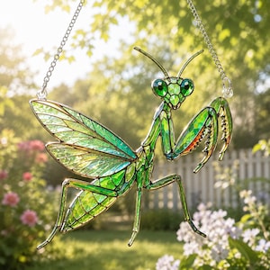 May include: A stained glass suncatcher shaped like a praying mantis, featuring vibrant green and iridescent glass segments held together by dark metal solder lines. The insect figure is suspended by a silver-toned metal chain attached to its top.