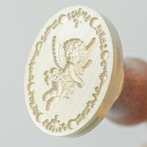 May include: A round brass wax seal stamp featuring an engraved cherub figure in the center, surrounded by a circular border of moon phases and small star motifs. The stamp is attached to a smooth, rounded wooden handle for easy gripping.