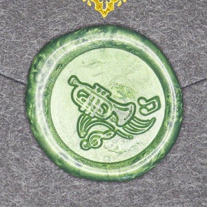 Puede incluir: Un sello de cera verde con un diseño de instrumento musical, que incluye una trompeta y notas musicales, está centrado en un sobre gris. Las palabras "Thank You" están escritas en oro encima del sello.